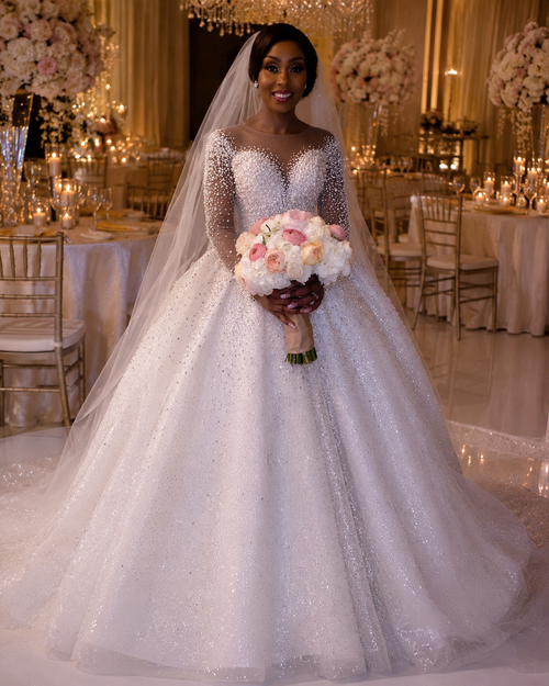 Ball gown wedding dress with long sleeves, adorned with pearls and a sparkly white skirt, paired with a bridal bouquet.